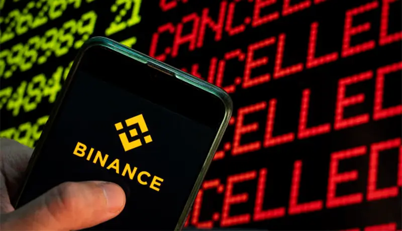 The logo of cryptocurrency exchange Binance displayed on a smartphone with the word cancelled on a computer screen in the background